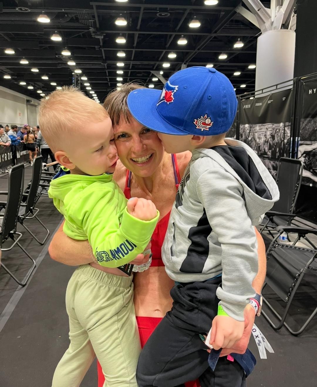 Woman holding two young children wearing blue Toronto Blue Jays baseball caps at an indoor event.